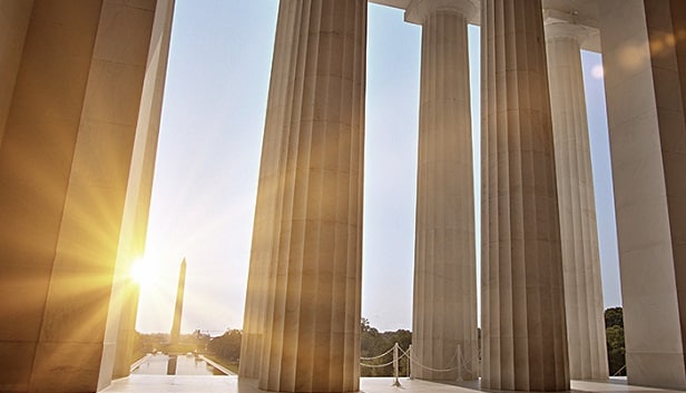 An image of the Washington Monument in the distance with the sun to the left of it, looking from the vantage point and framed by the columns of the Lincoln Memorial