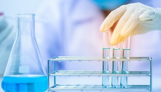 A close-up image of a lab table with a beaker to the left, and a hand wearing a protective glove holding 3 test tubes 2/3s filled with clear liquid to the right