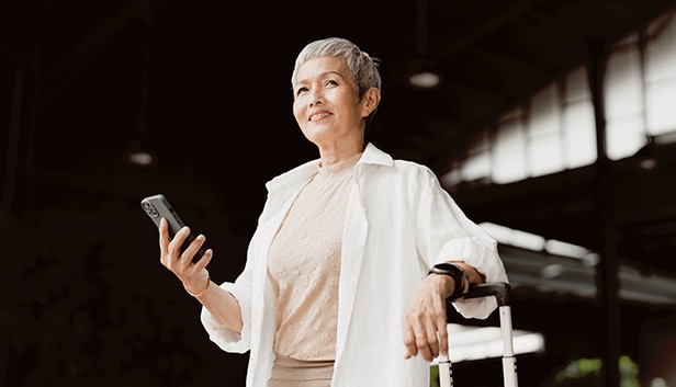 An image of a gray-haired woman looking at her cell phone in one hand with the other hand on the handle of her carry-on luggage.